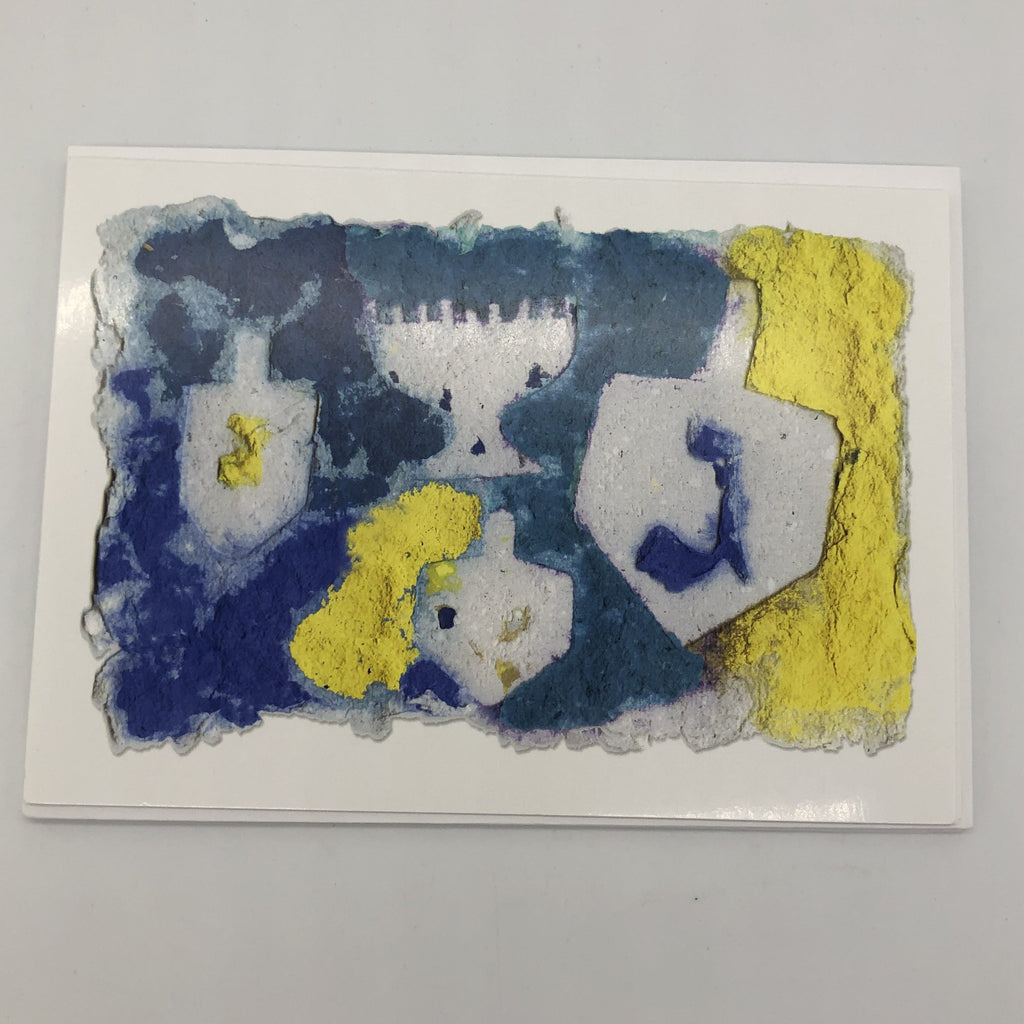 Printed card of a photograph of handmade paper in blue and yellow with four white elements on top.  The elements are two small dreidels, a large dreidel with the Gimmel symbol and a menorah image, all in white.