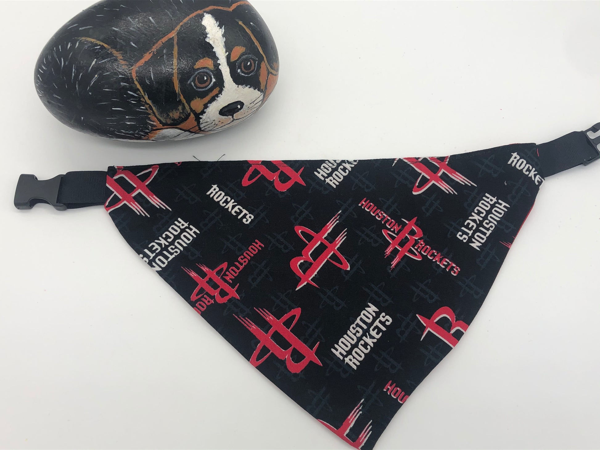 Decorative rock painted like a small curled up black and tan dog next to a blak Houston Rockets triangular dog bandana with black plastic closure.  Bandana is primarily black with red Rockets logo and Houston Rockets written in white.