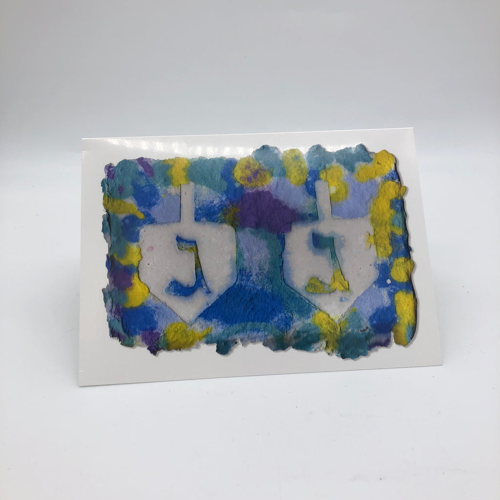 White greeting card with the printed image of handmade paper in shades of yellow, blue and a little bit of purple.  There are two large dreidels side by side with the Hebrew symbol 