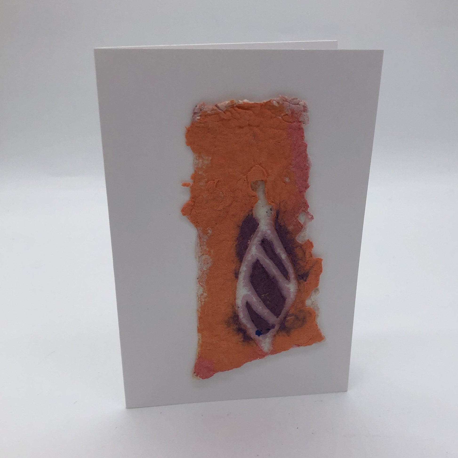 Handmade paper card made with orange background with white pointy oval ornament on top,  Ornament is purple with white stripes.ornament