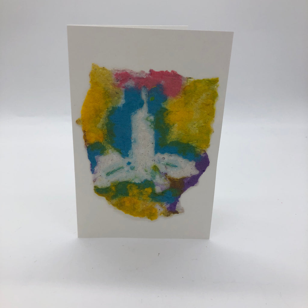 Handmade paper candle with multiple colors in the background including teal, yellow and pink.  White candle with white holly around it.