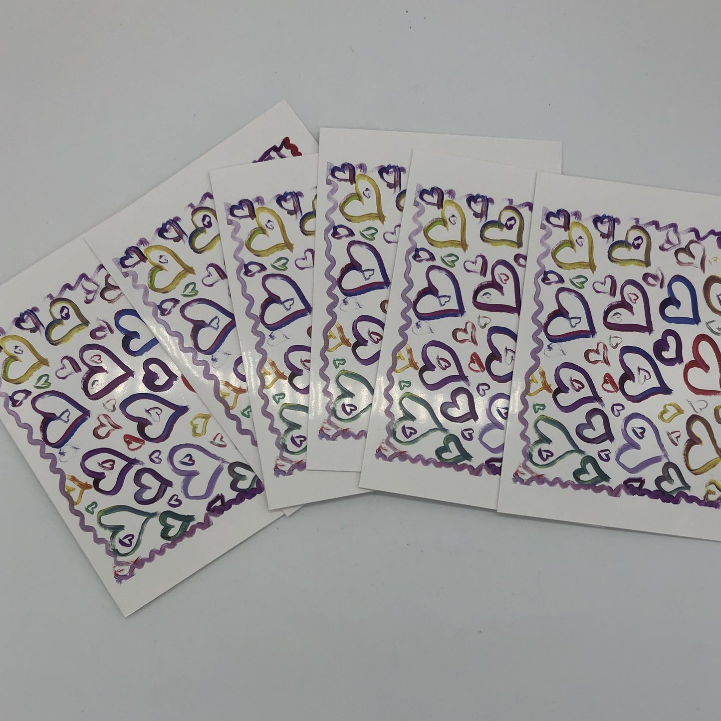 Six of the "Hearts" greeting cards fanned out. White greeting card with about a dozen large water colored hearts in blues, purples, and yellows. There are several smaller hearts of varying sizes all around those larger hearts and a squiggly line in shades of purple and red framing all of it.