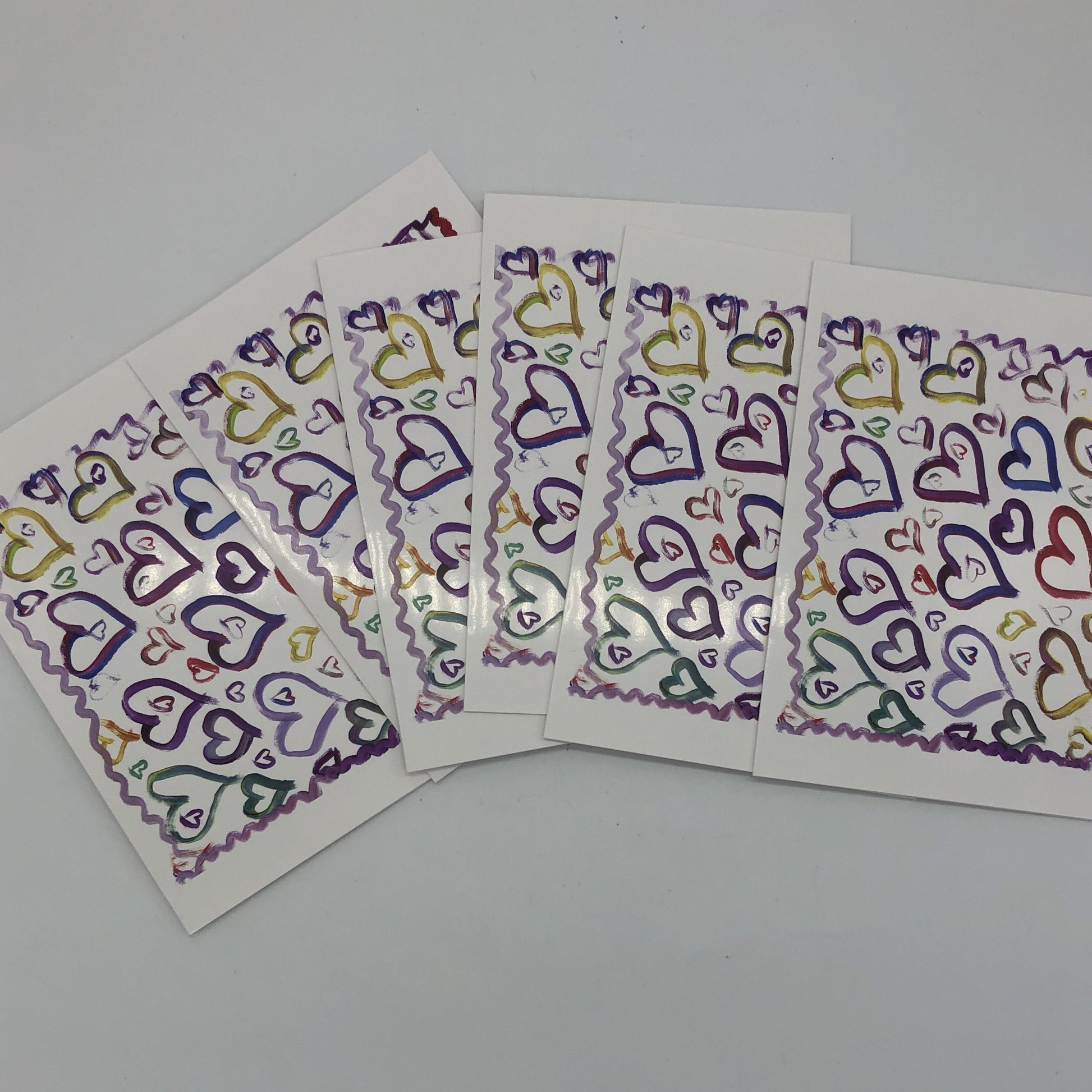 Six of the "Hearts" greeting cards fanned out. White greeting card with about a dozen large water colored hearts in blues, purples, and yellows. There are several smaller hearts of varying sizes all around those larger hearts and a squiggly line in shades of purple and red framing all of it.