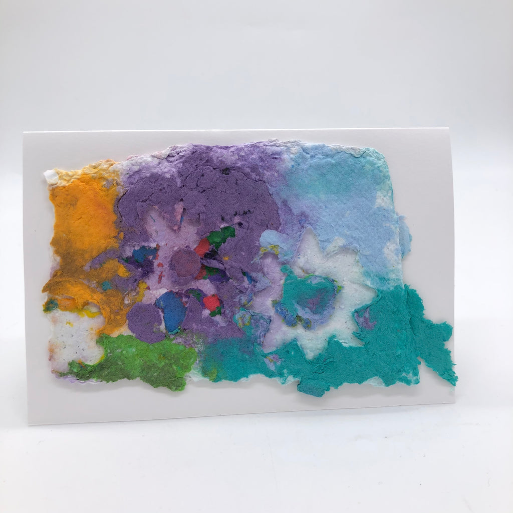 Handmade paper greeting card with background in pastel shades of purple, blue, teal and green with bright yellow.  On top are two floral shapes, one with several small dots of color, the other with white petals and a teal center.
