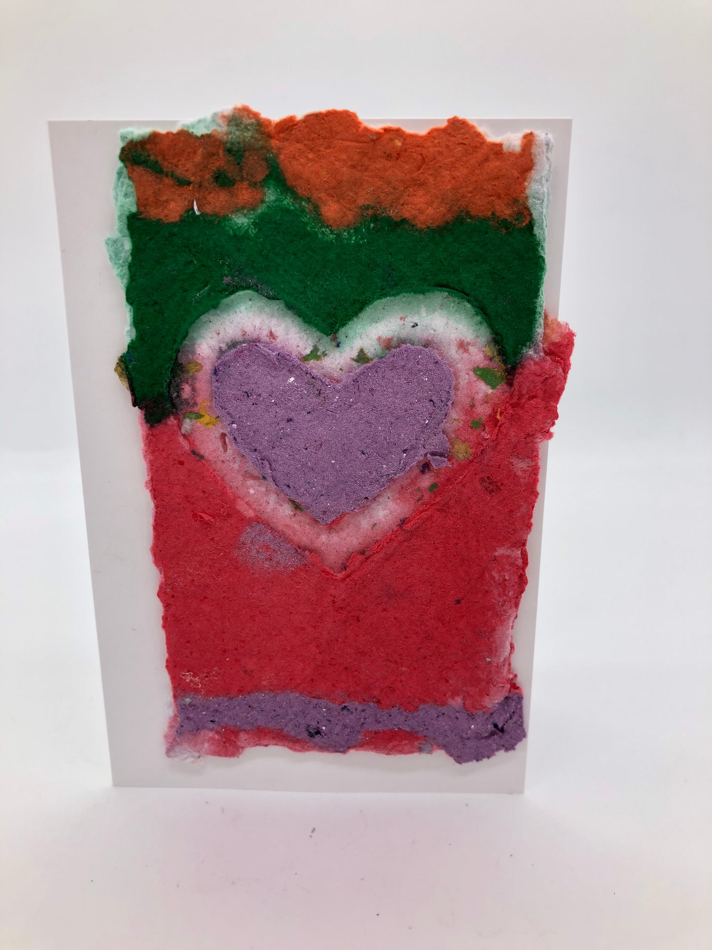 Handmade paper greeting card with a white outline heart.  Inside the heart is lavender.  The background has horizontal stripes of color with orange at the top, then dark green, red from center to the bottow with a think purple stripe at bottom.  