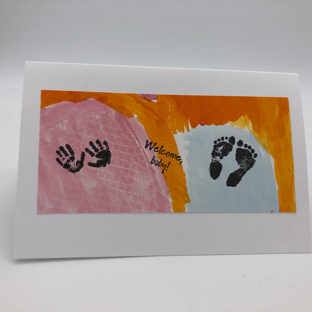 Hand painted greeting card in pink, orange and light blue.  On top of color are stamps, to the left hand side is two small handprints in black.  In the middle it says Welcome, baby! at an angle and in black and to the right side are two tiny black feet.