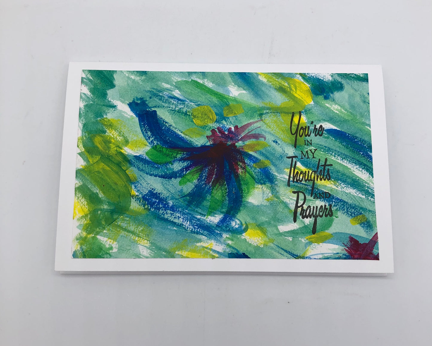 White greeting card with large acrylic painting in shades of greens, blues and yellow.  Some of the white background is showing through.  In the center ti a partial swirl in blue..  On top of the painting is stamped "You're in my thoughts and prayers" in black on the right hand side.