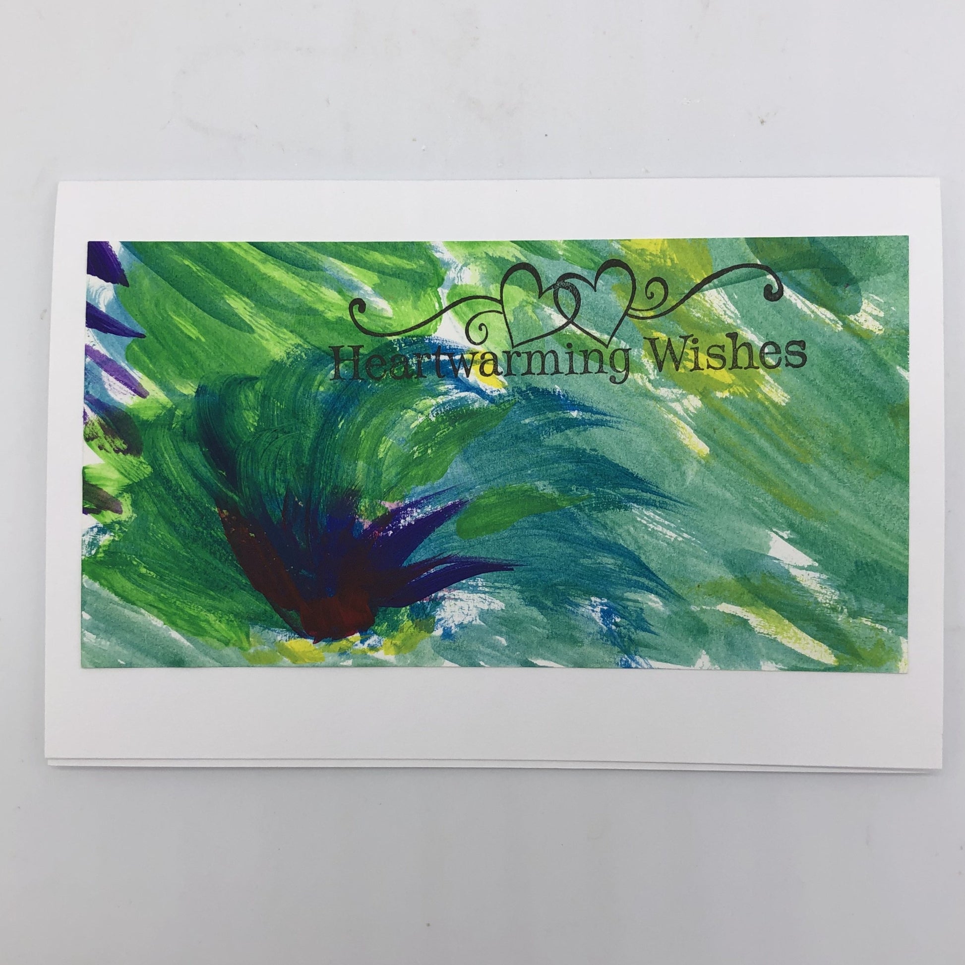 White greeting card with shade of blue and green painted on top is lines.  There is a dark blue cluster resembling a tuft of grass in the lower left hand corner.  On the top portion of the painting, flush to the right is the stamp in black that says Heartwarming Wishes and has two outlined hearts touching with flourishes to each side.