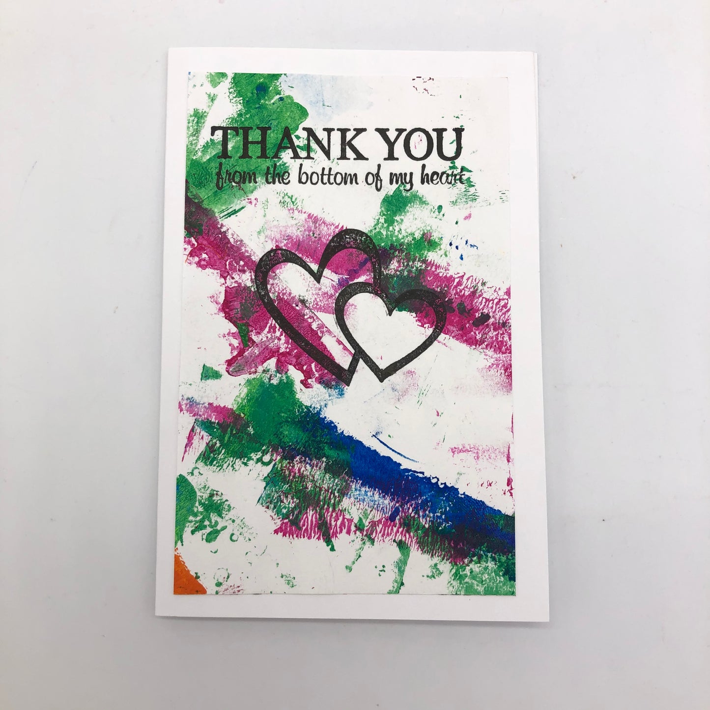 White greeting card with paint strokes of green, maroon and blue.  Quite a bit of the white is showing through.  In the middle is are two stamped outlined hearts, one larger than the other.  At the top is says THANK YOU from the bottom of my heart.