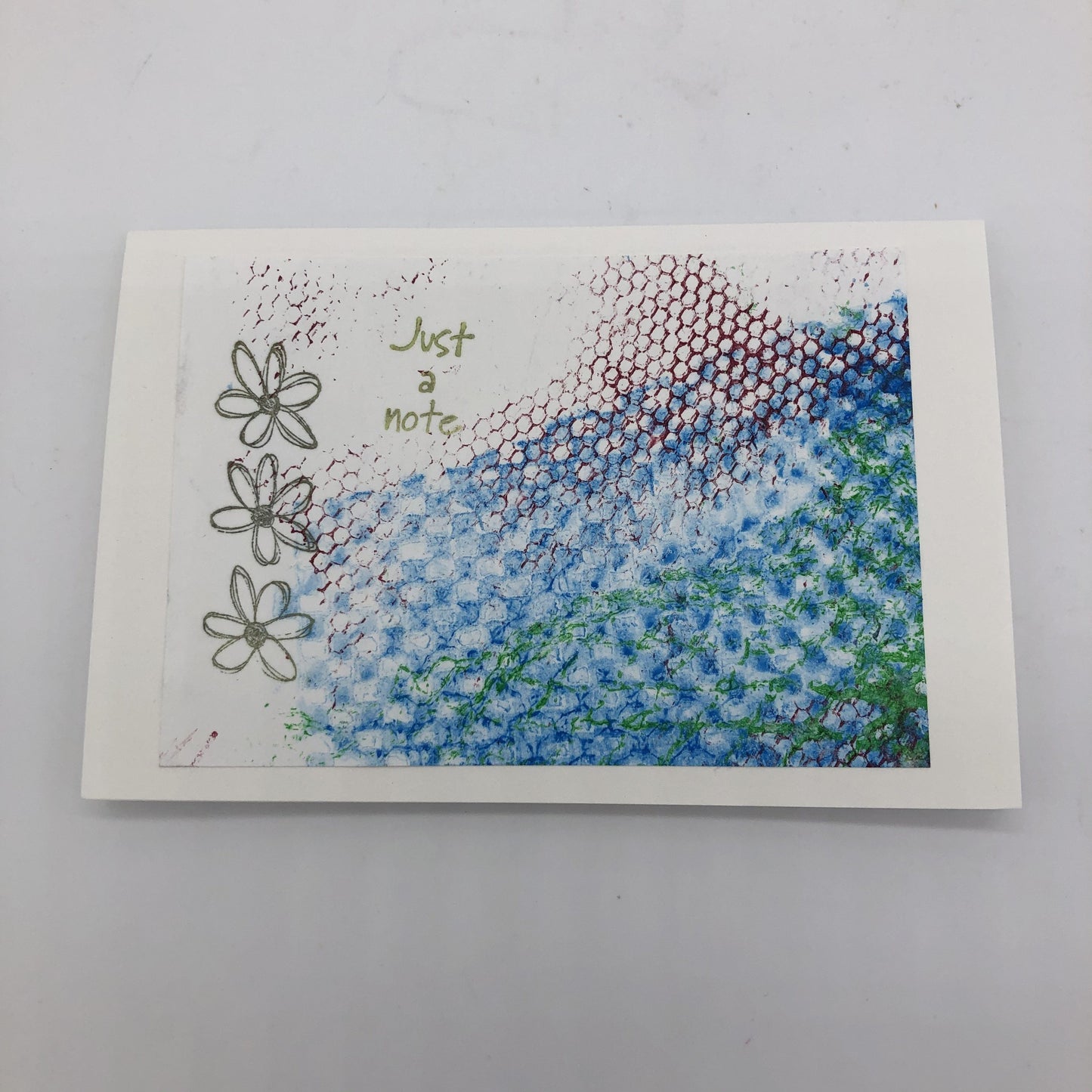 White greeting card with rubbings of a textured net material in green and maroon.  There is also a large swatch of lined up circle texture in blue.  On top of the texture are three stamped outlined daisys in black and the words "Just a note" in green.