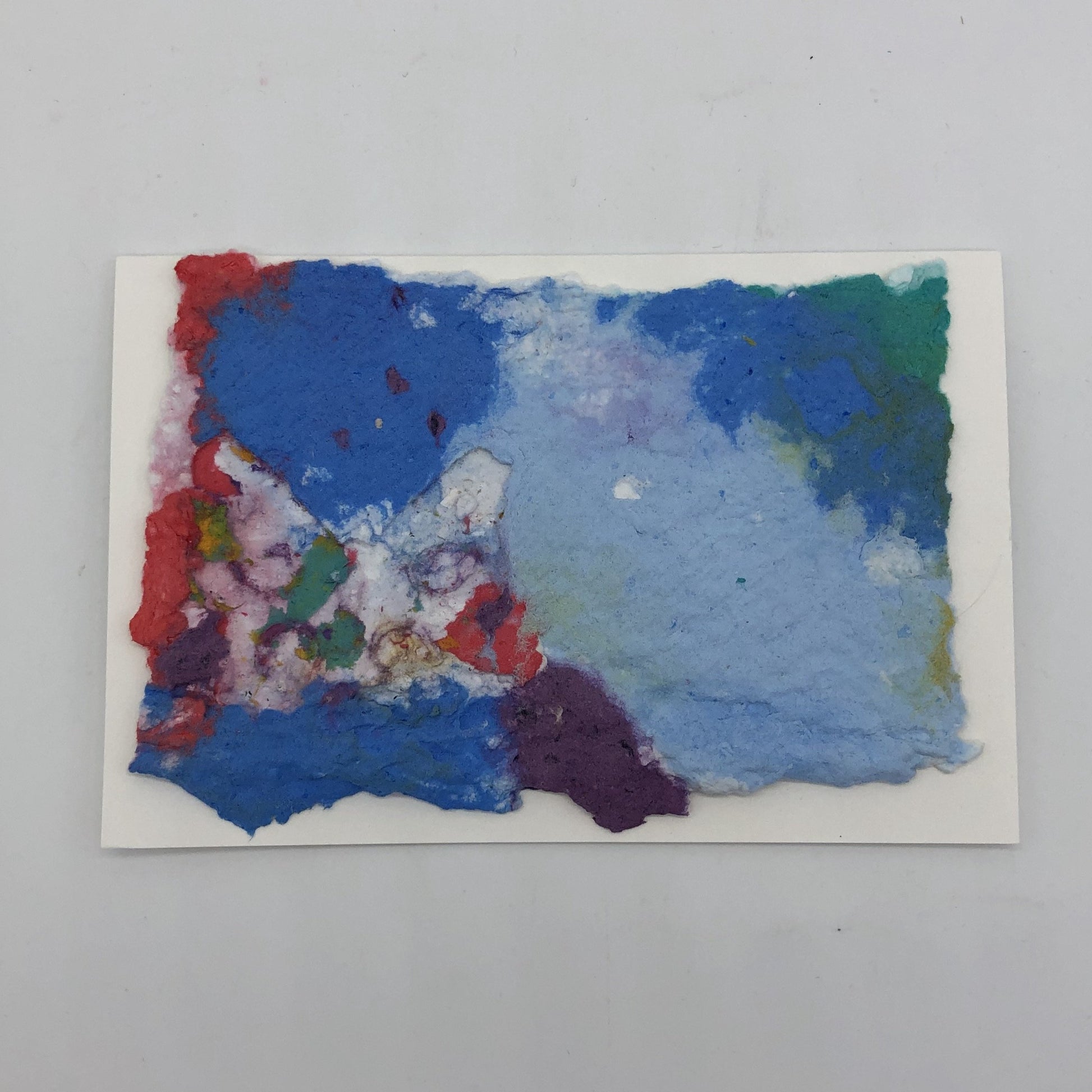 Handmade paper greeting card of blues, purple , red and green colors melted together in the background with a white small butterfly over top int the lower left hand corner