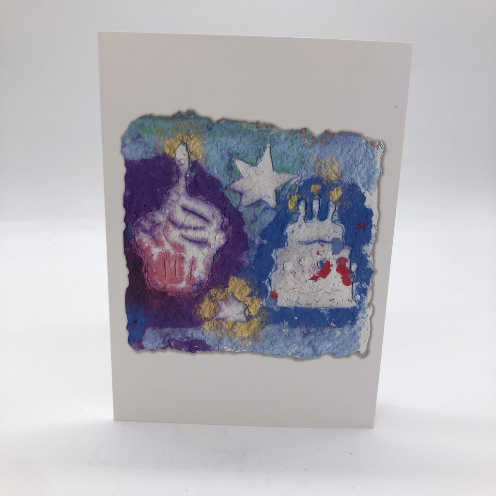 Printed card of a picture of handmade paper in blues and purple with a little gold.  On top of the paper are stars, cupcake and cake with candles.