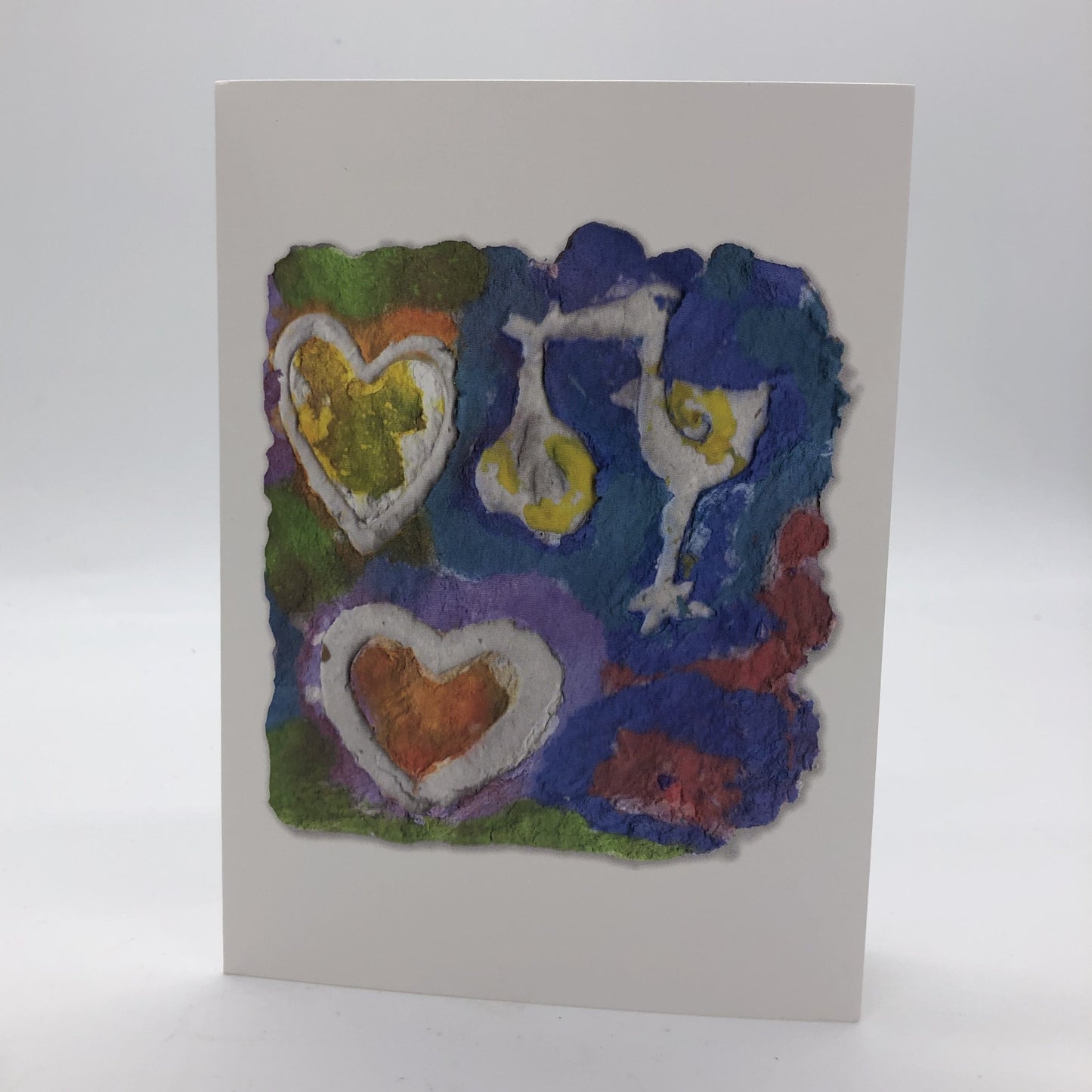 Printed card of photo of handmade paper in blues, greens and reds. On top of the design are two white hearts and a stork holding a wrapped baby by it's beak.