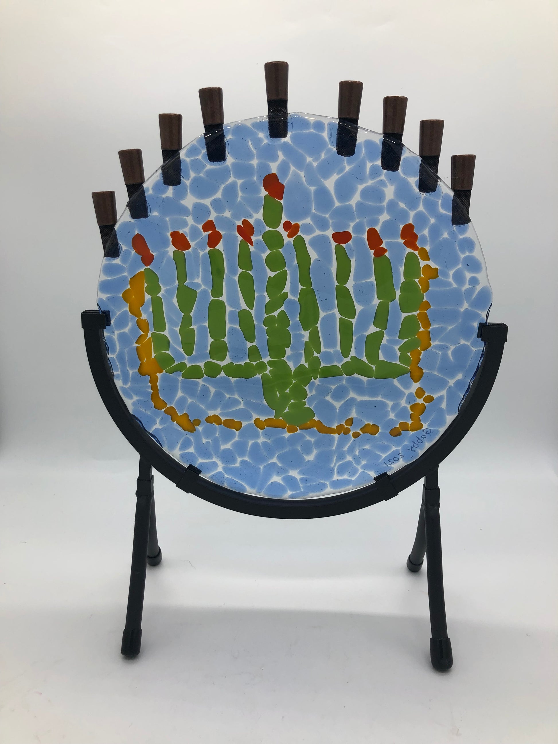 Round fused glass piece with design of a menorah done in green with yellow outline around three sides.  the menorah is surrounded by blue chunks of glass.  There are nine candle cups on the top of the glass and is held in by a semi-circular metal frame.