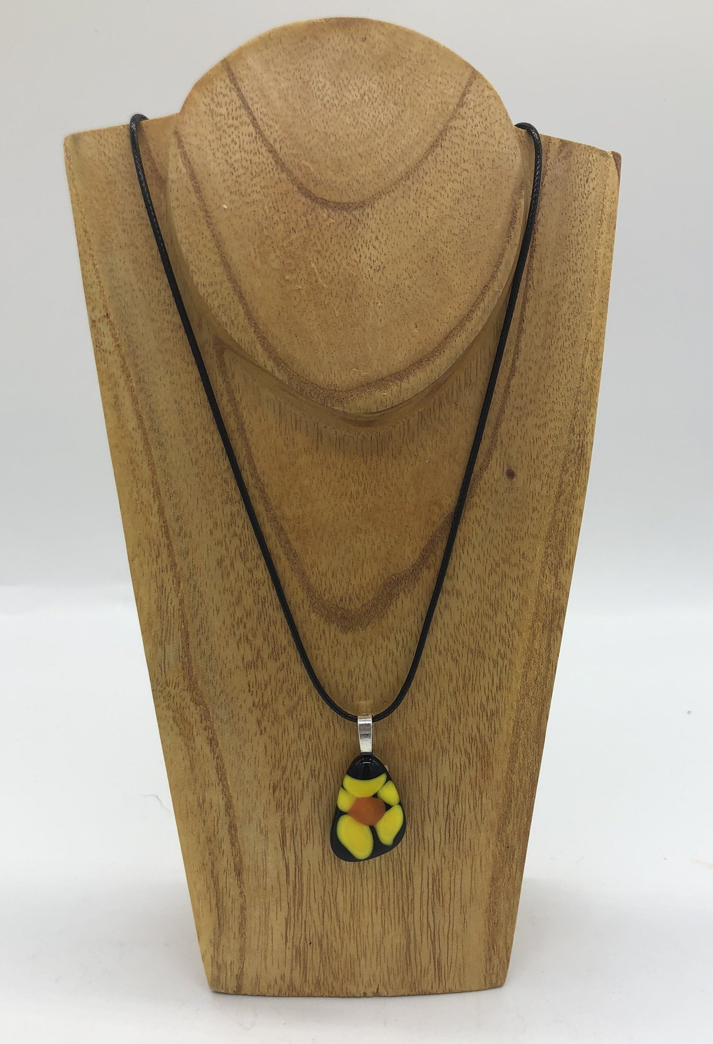 Fused glass necklace on a black cord, displaced on wooden holder.  Necklace design is a black oval with yellow petaled flower with orange center.