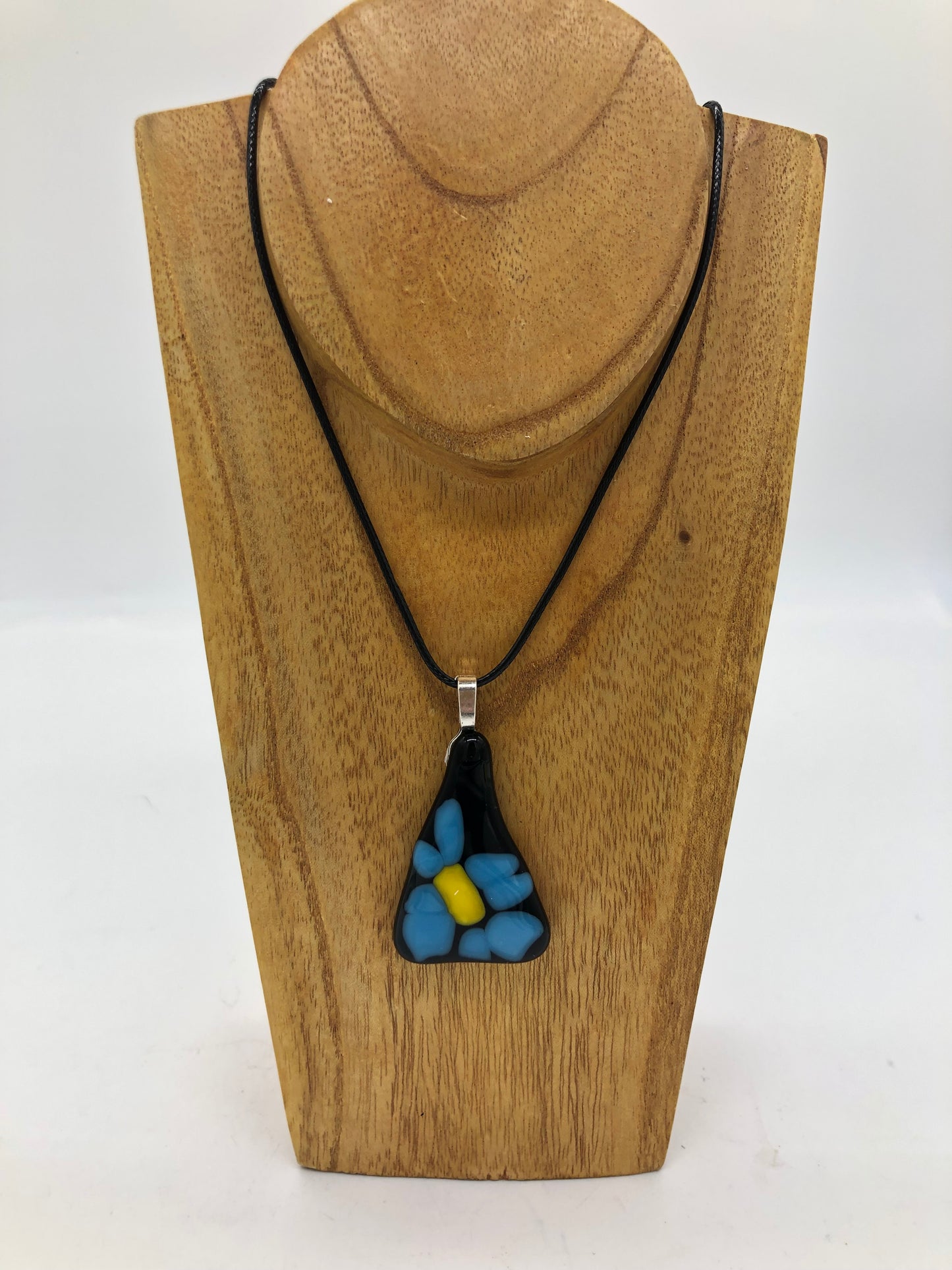 Fused glass necklace on a black cord, displaced on wooden holder.  Necklace design is a black triangle with blue petaled flower with yellow center.