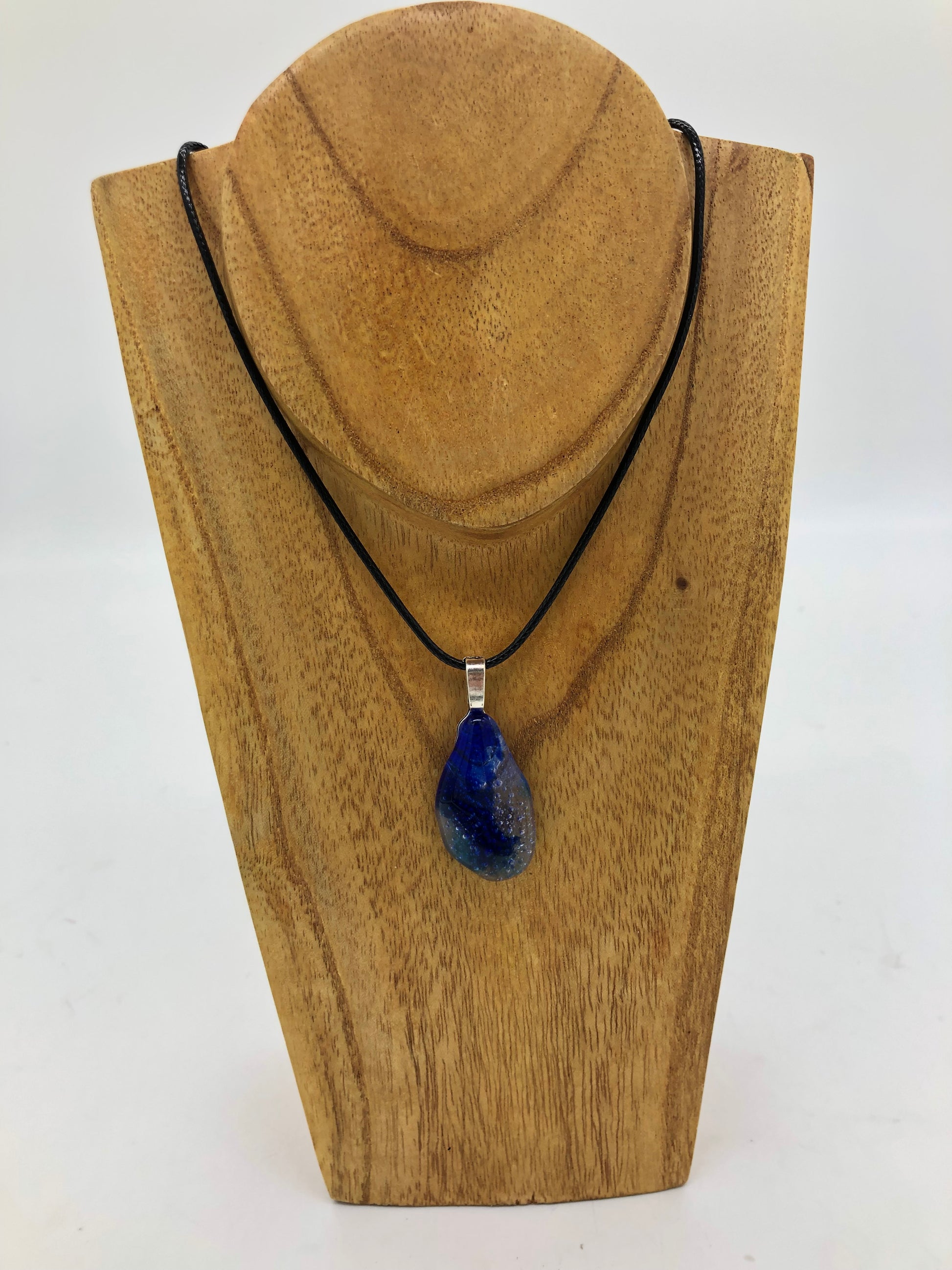 Fused glass necklace on a black cord, displayed on wooden holder.  Necklace design is a elongated freeform oval in blues and clear