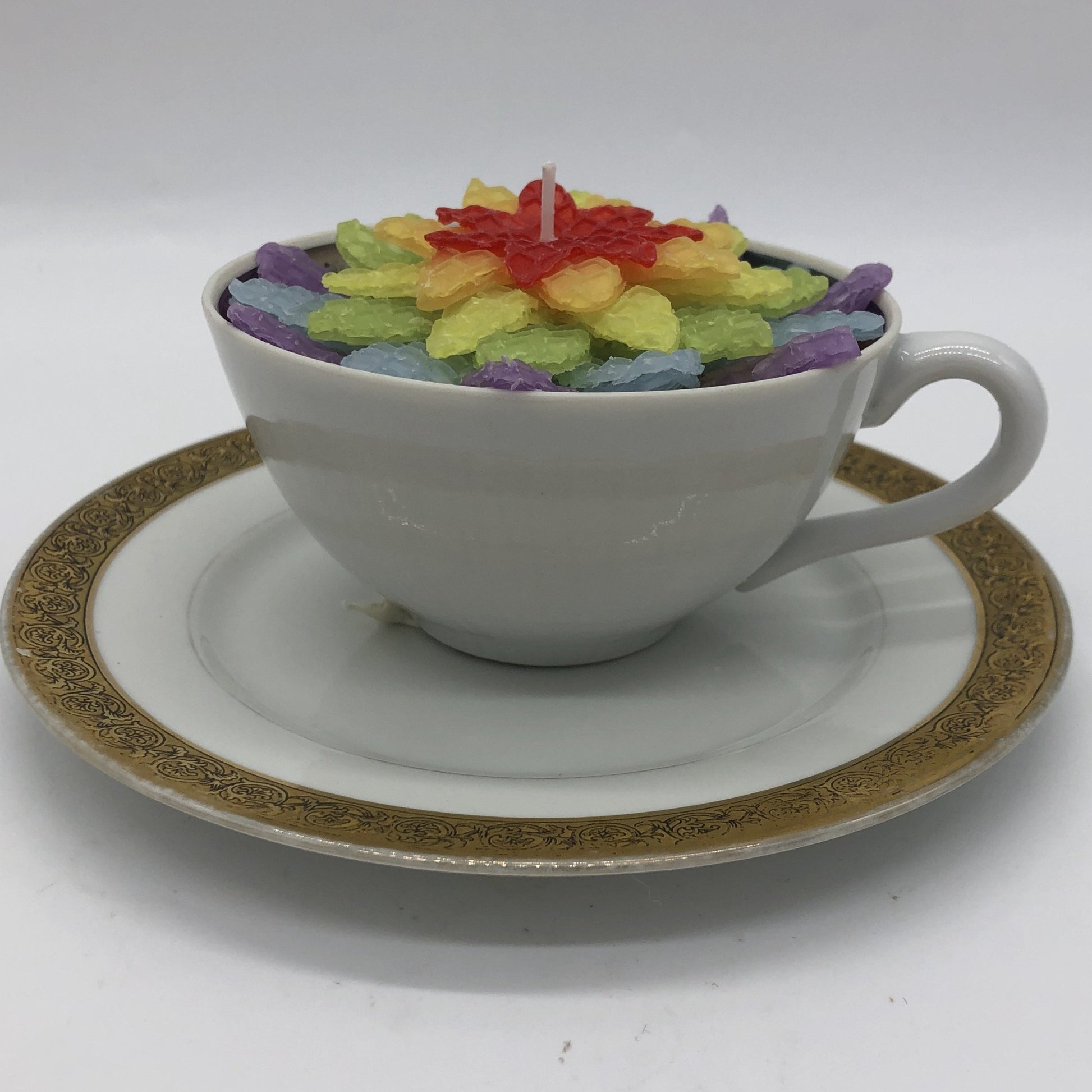 White teacup with white saucer with fancy gold border.  Inside teacup in a six layer flower in the colors of the rainbow.