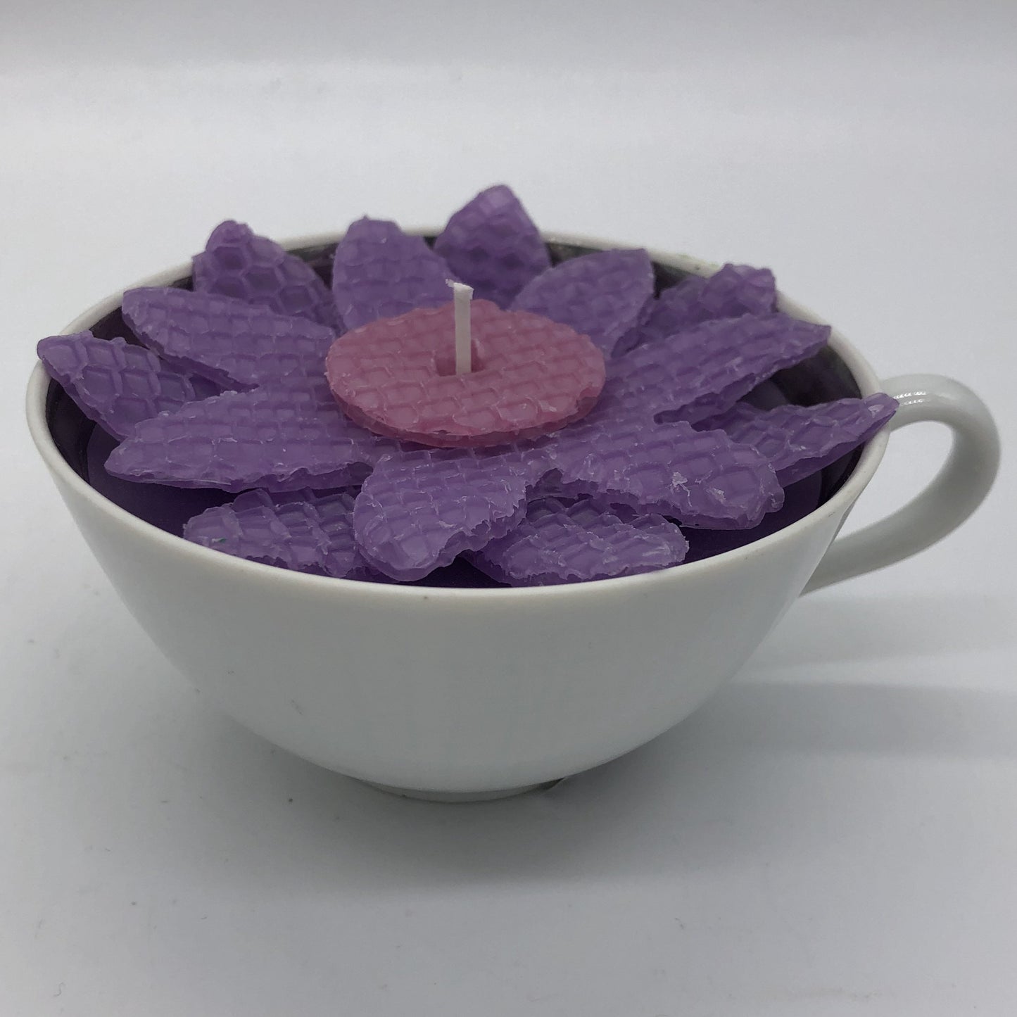 White teacup filled with candle and purple and pink daisy made from beeswax sheet on top.