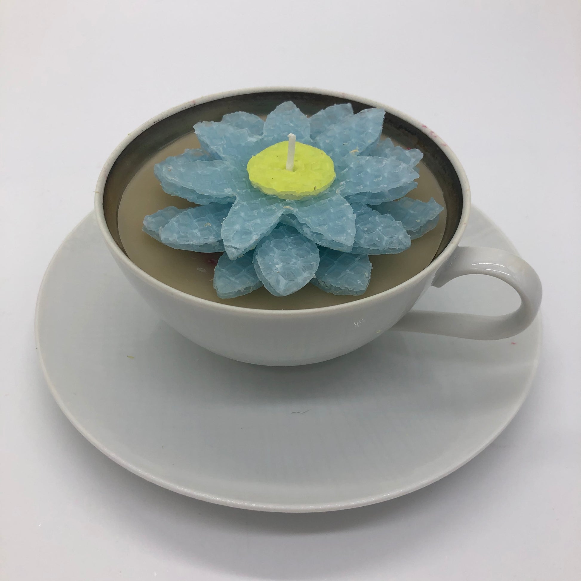 All white teacup and saucer with light colored candle inside.  Top of candle is decorated with a multilayer light blue flower with yellow center made of beeswax sheets.