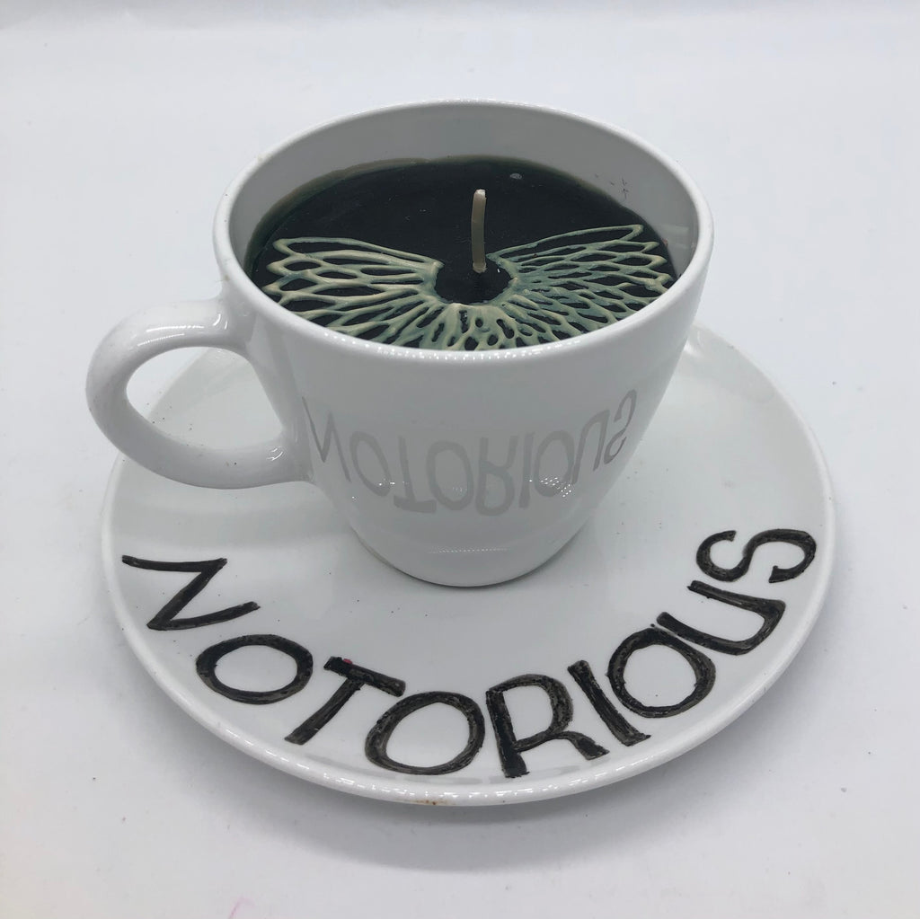 Solid white teacup and saucer with black candle inside.  On top of candle it has a lace collar.  The saucer says NOTORIOUS.