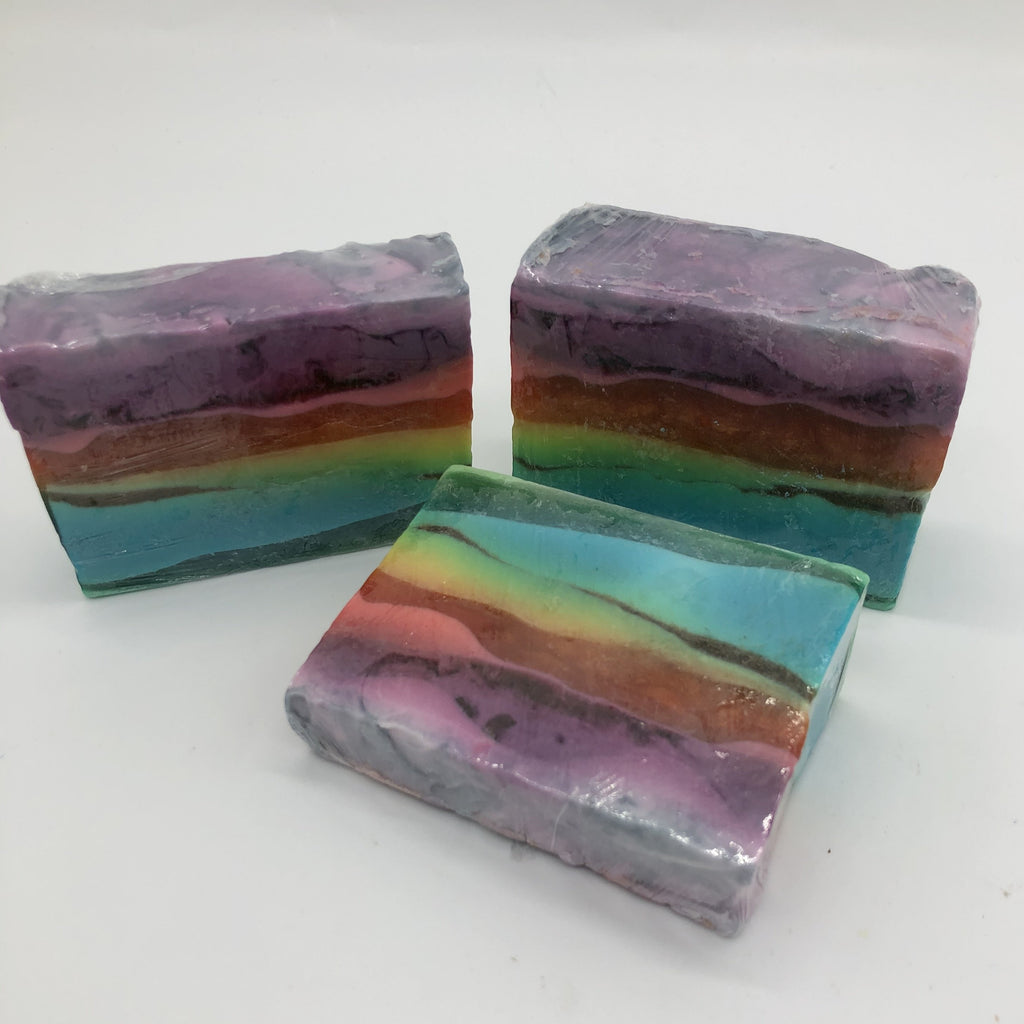 Three bars of rectangular soap with several layers of color including creamy plum, translucent purple,yellow, orange, creamy aqua and translucent green. Two bars are sitting on the bottom edge, one in front is laying down.