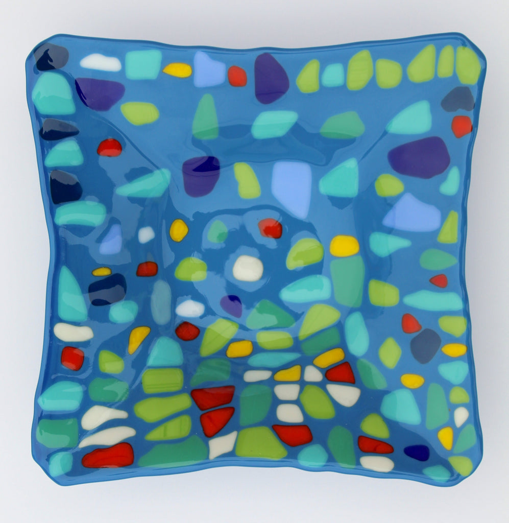 This is a sqaure fused glass piece with a blue background. There are pale blue, green, dark blue, red, yellow and white shapes over it