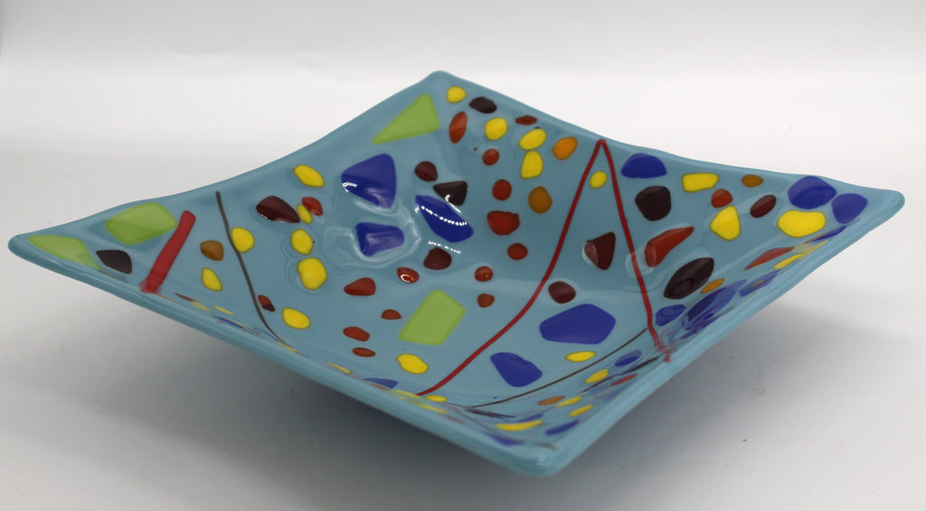 This is a blue sqaure glass bowl with red lines across the middle. It also has several abstract shapes in green, yellow, black, brown and blue