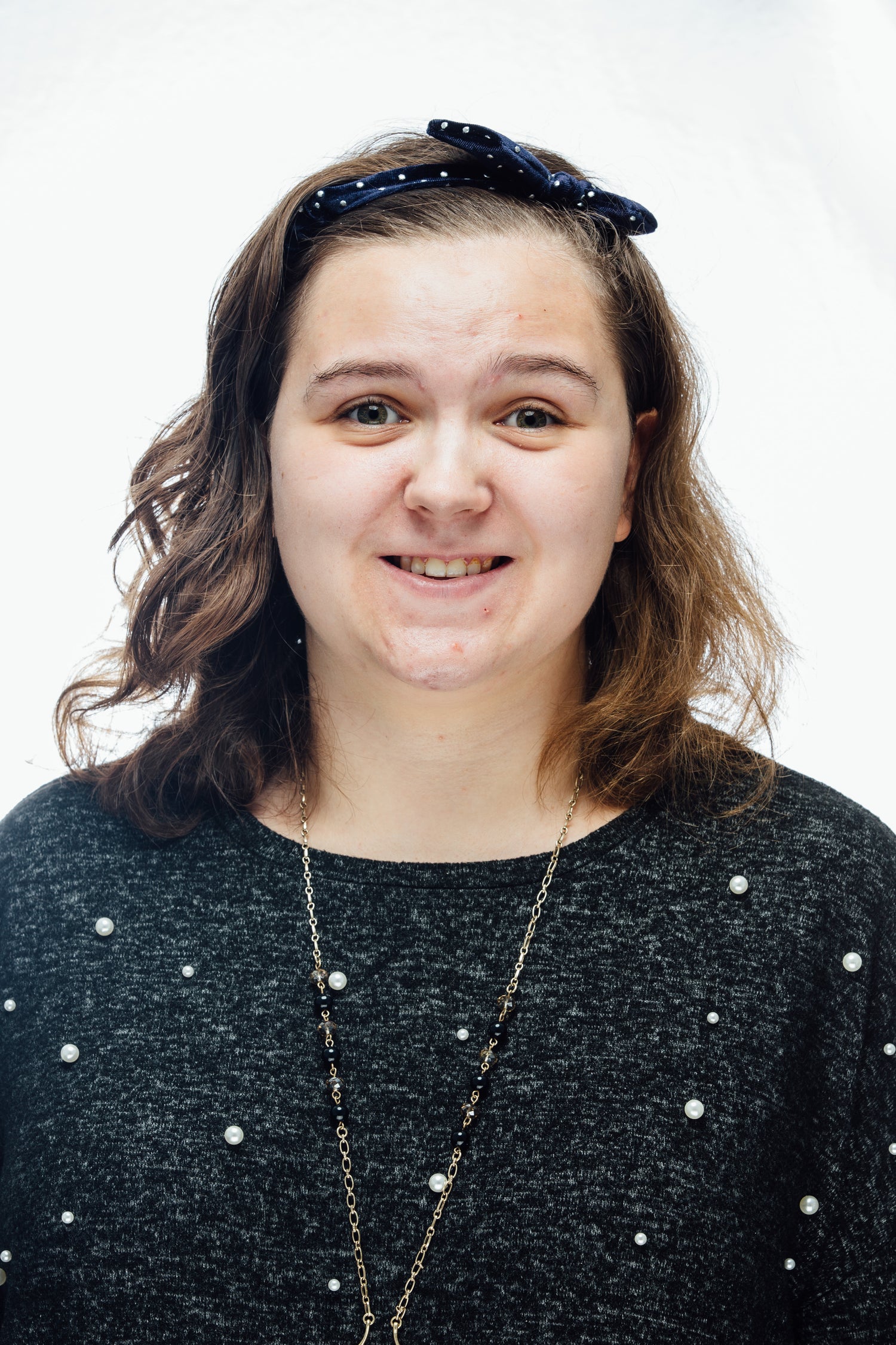 Smiling young woman with brown hair wearing blue bow headband and black sweatshirt with pearls