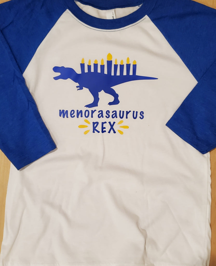 White tshirt with blue sleeves with a T-Rex dinosaur with a menorah on its back with blue lettering spelling Menorasaurus Rex below
