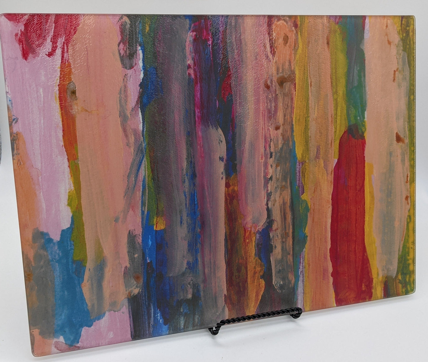 Large Rectangle glass cutting board with the following image of Layers of vertical brushstrokes overlapping. In the center the background is a dark blue, to the left is yellow, and the right is pink. The top layer of brushstrokes are mostly a pale pink