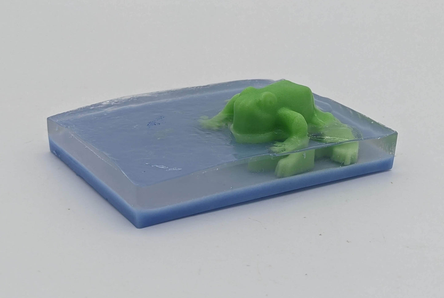 Bar soap with a green frog protruding. There is a thin layer of blue, then clear, and the green frog is embedded in the clear portion to appear as is the frog is sitting on top of water