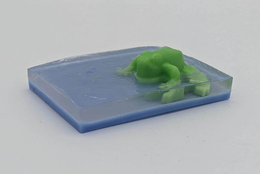 Bar soap with a green frog protruding. There is a thin layer of blue, then clear, and the green frog is embedded in the clear portion to appear as is the frog is sitting on top of water