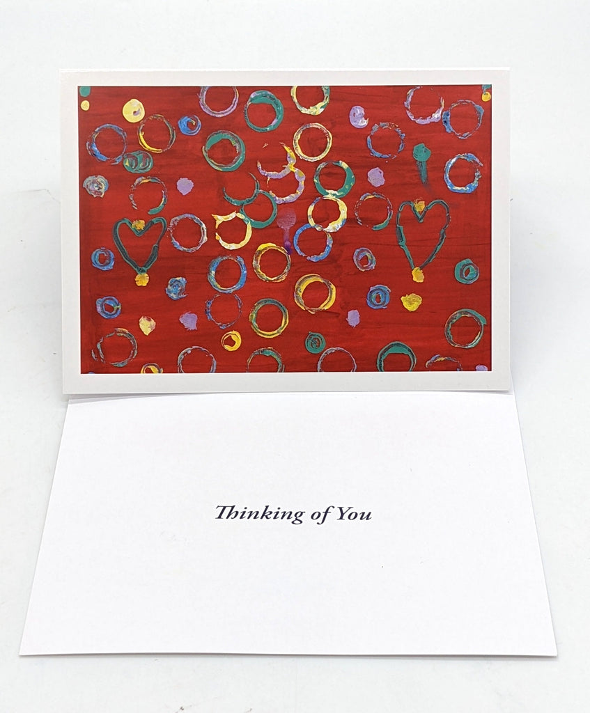 This is a card that says thinking of you. It has a painting with a red background. It has several blue, pink, and yellow circles and two green hearts
