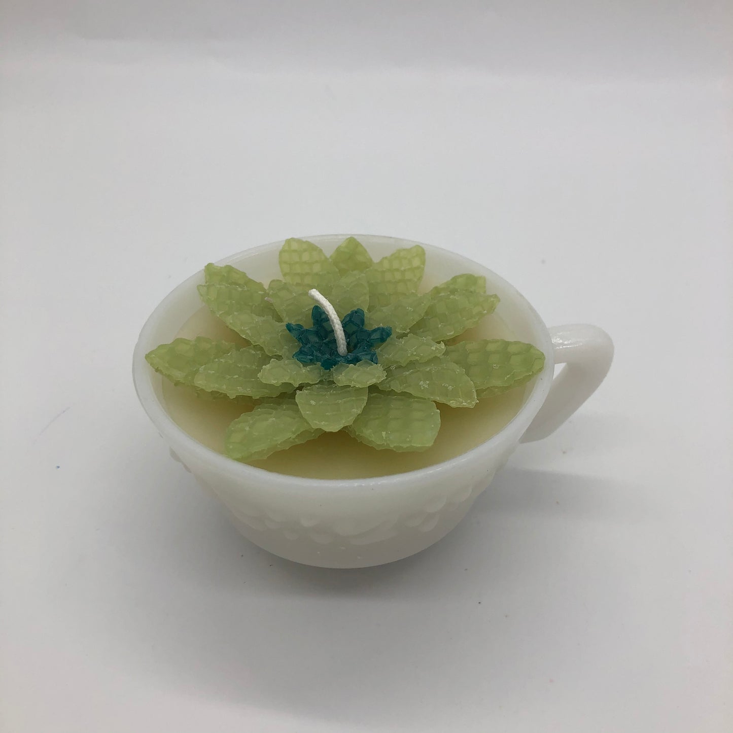 Milk glass teacup with light yellow candle and light green beeswax sheet succulent with small darkgreen centeron top.
