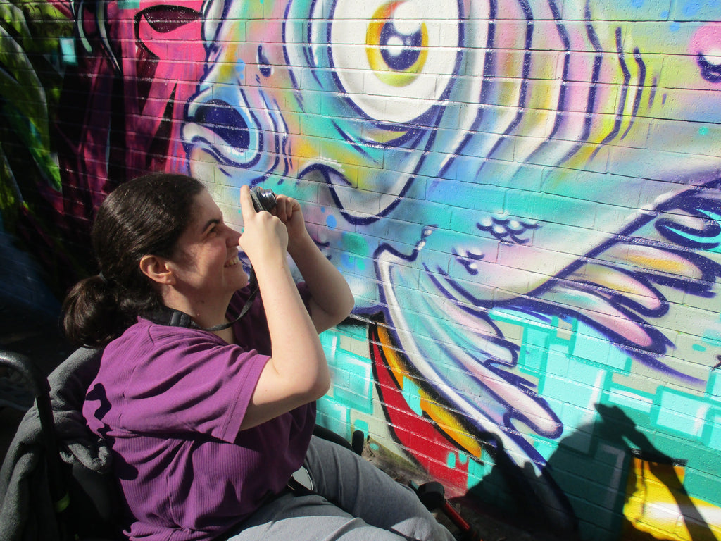 Female participant taking a photo outside.   She is sitting in her wheelchair, has her hair pulled back  and wearing a purple t-shirt.  The sugject of her photograph is a colorful fish mural on a brick wall.
