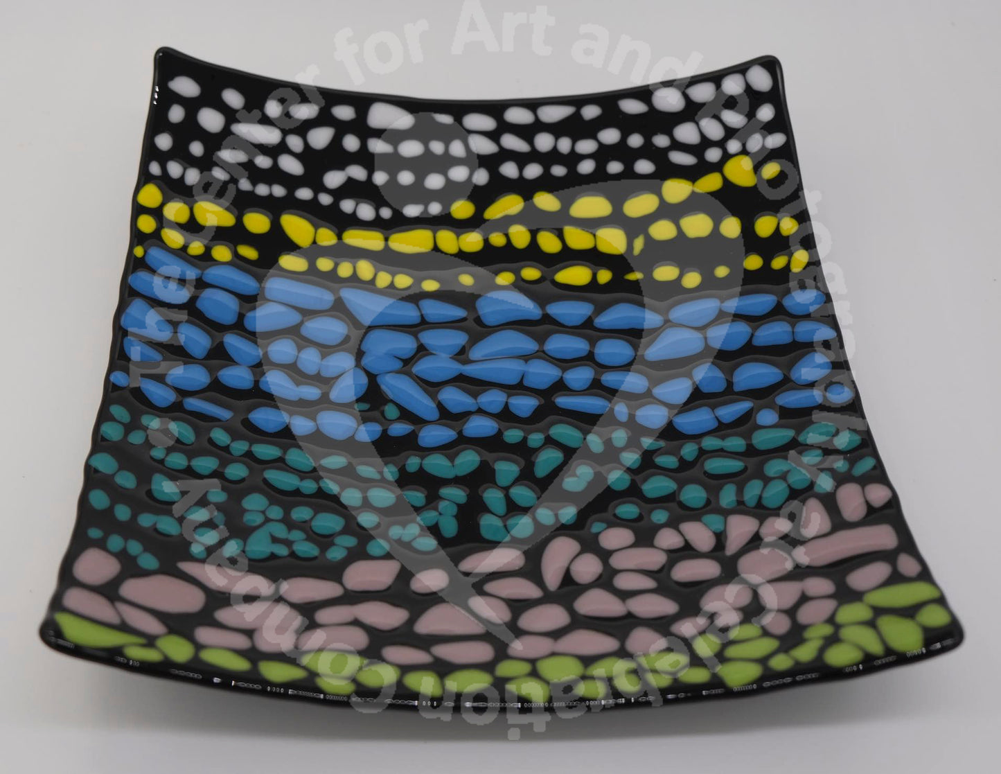A black square glass curved plate that has small pebbles of colors. There are on average of three rows each color before transitioning to the next color. Colored pebbles are placed in the following order: white, yellow, blue, turquoise, light purple, and green.