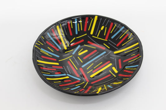 This is a black circular glass bowl with yellow, blue, and red lines in an abstract pattern