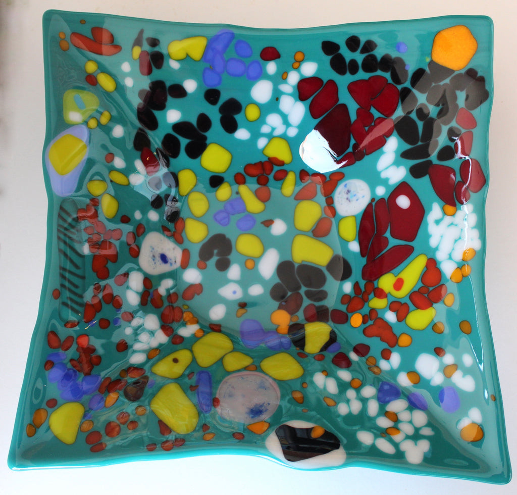 This is a fused glass piece with a blue background and several abstract shapes including: yellow, red, white, orane, black, and blue