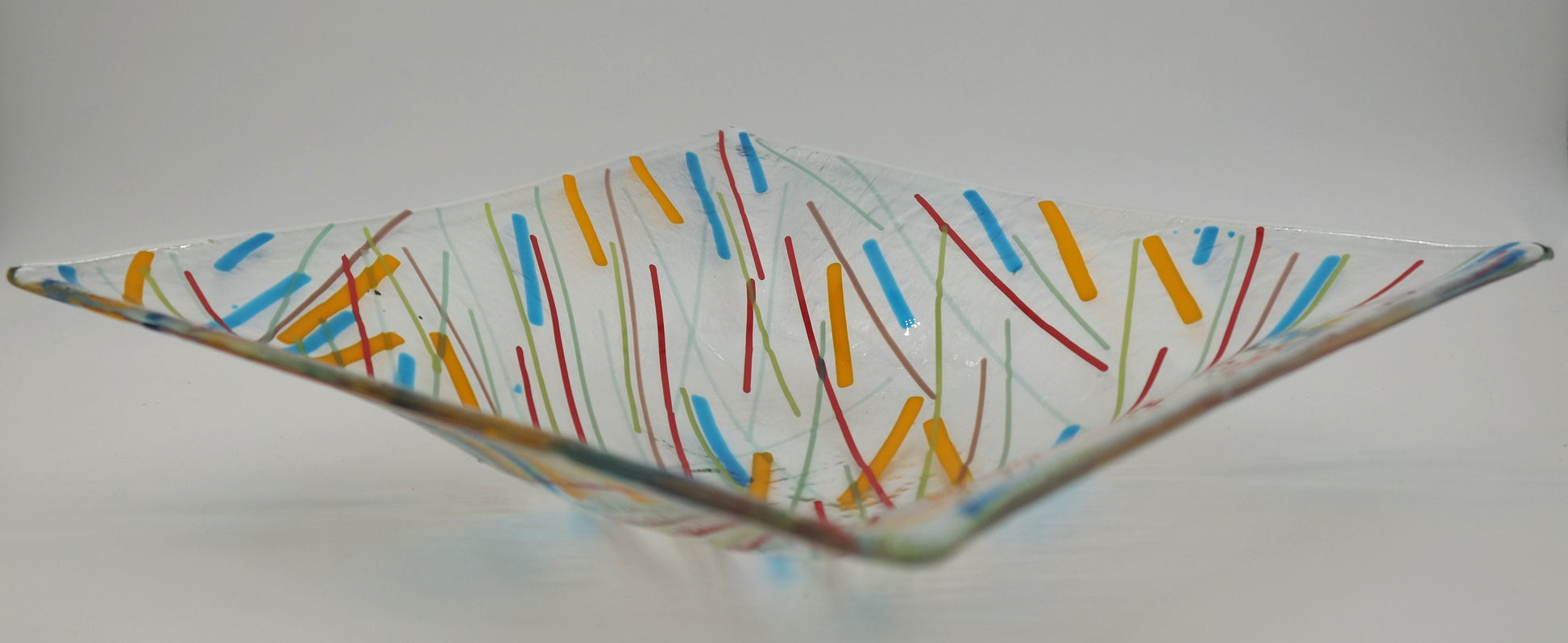 Side view of a Fused glass square bowl. Bowl is a made of a clear piece of glass with lines radiating from the lower left corner outward. The colors of the lines are red, green, faint blue, with thicker lines of orange and light blue.