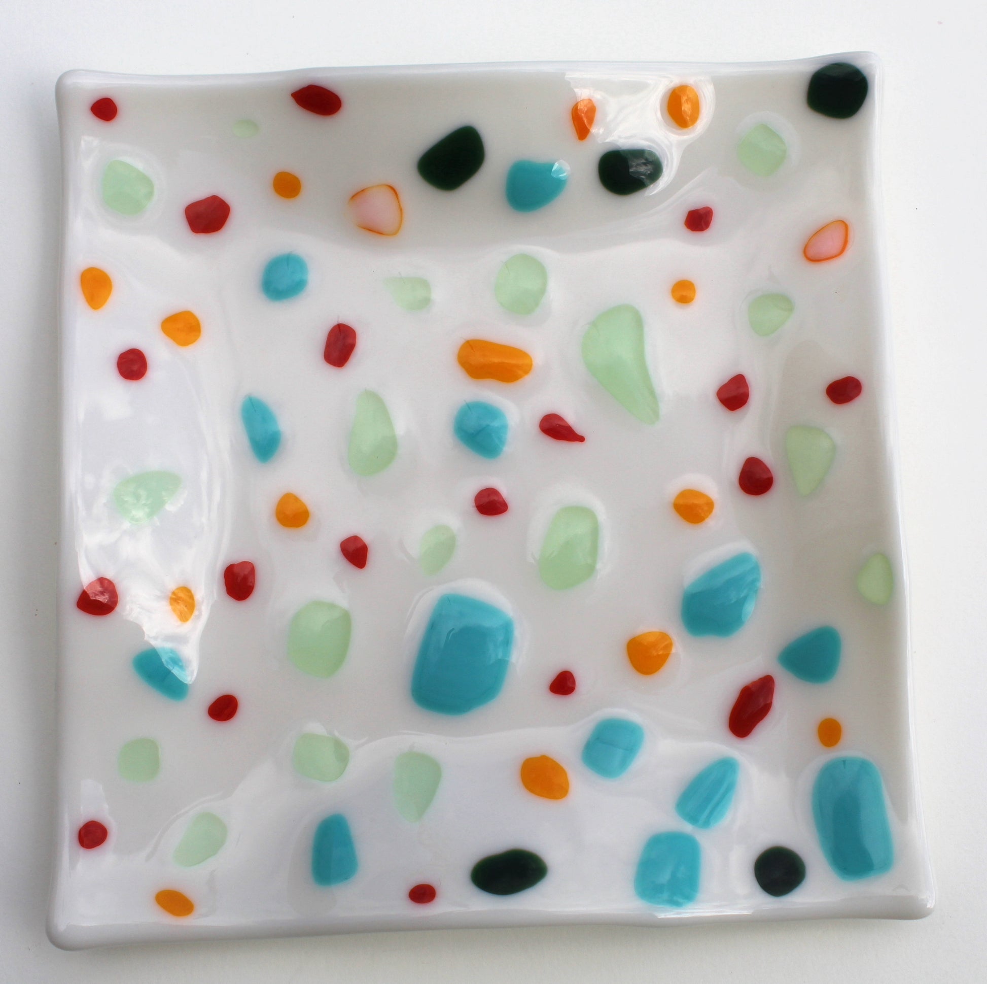 This is a white glass art piece in the shape of a square. It has multicolored blotches of blue, pale green, red, black, and orange,