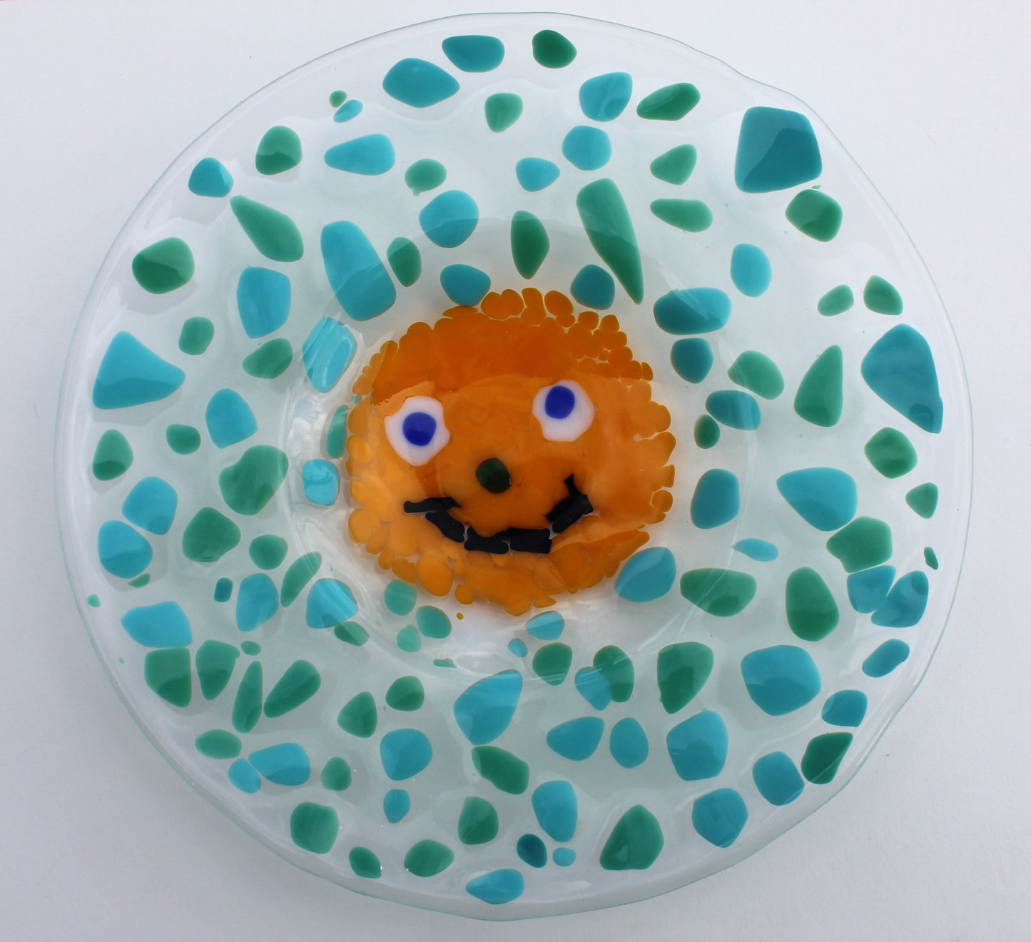 This is a round glass art piece with blue and green splothces around the 4dges. In the center there is a yellow smiley face with a black smile and blue eyes