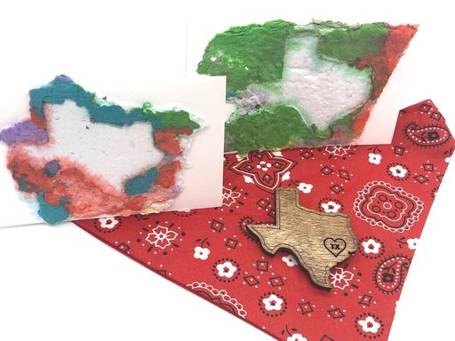 Two handmade paper state of TExas cards on top of a red bandana.  There is also a small wooden state of Texas cutout with a heart and TX on the wooden heart.