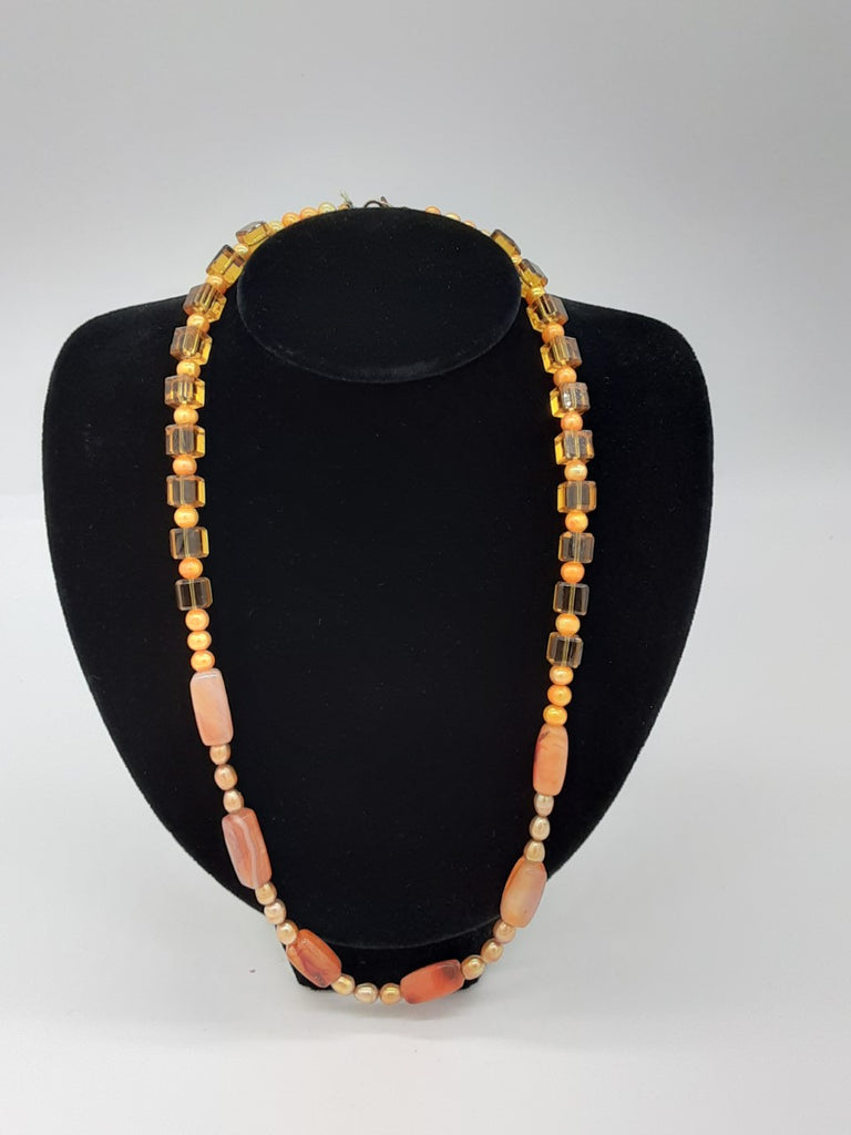 Orange necklace with a variety of bead shapes. Around the neck- clear yellow/orange cubes  and round opaque beads. In the center- gold glass oval pearls and cylinders of agate