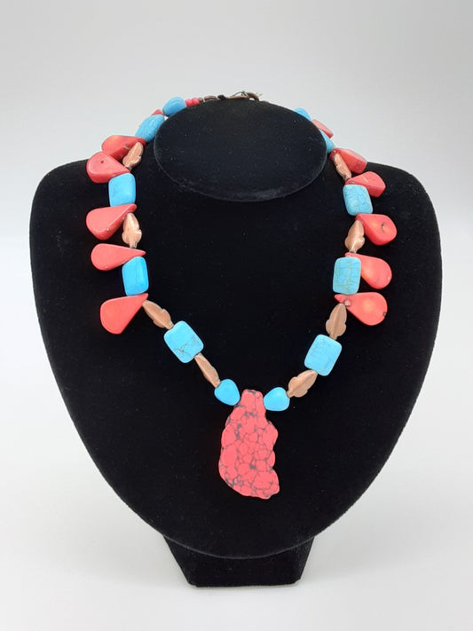 Necklace alternating in turquoise square beads, coral flat tear dropped shaped beads, copper shaped leaves. There is a large center pendant of an irregular cut red and black stone