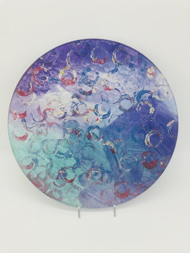 This is a round cutting board with the following painting: This is a painting with a green and purple background and circular imprints on top in red white and blue